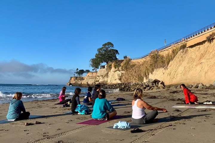 Group practicing yoga on a beach near cliffs under a blue sky.