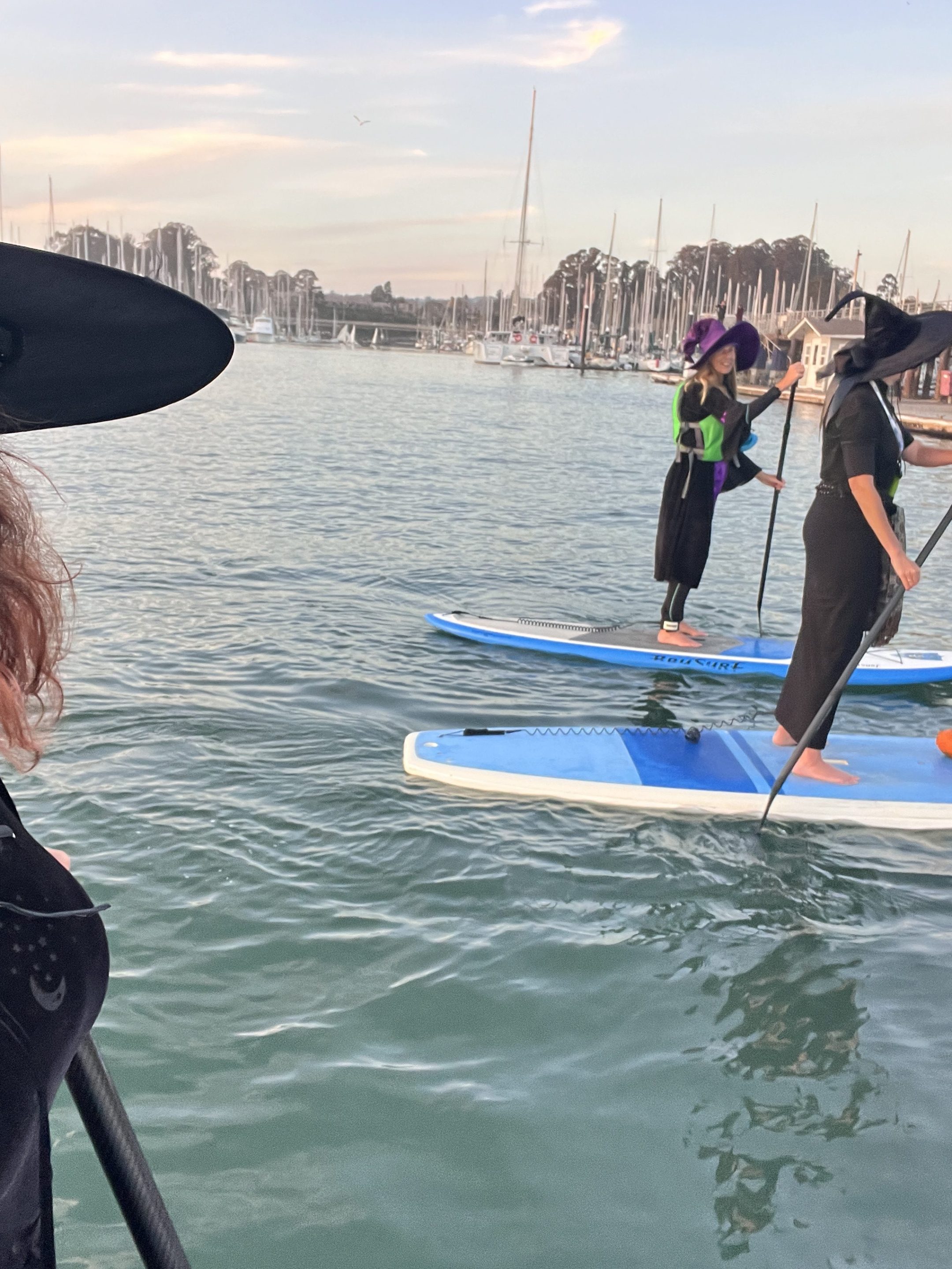 Woman in hat taking selfie, two people paddleboarding on calm water, sailboats in background.