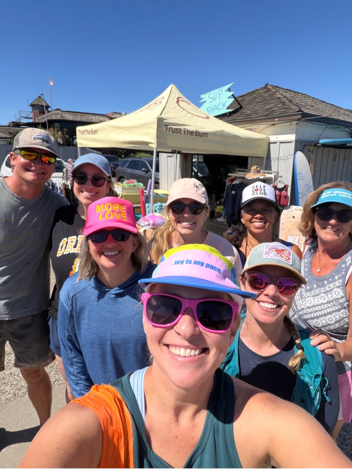 Group of seven people smiling outdoors, wearing hats and sunglasses, with a beige tent in the background.